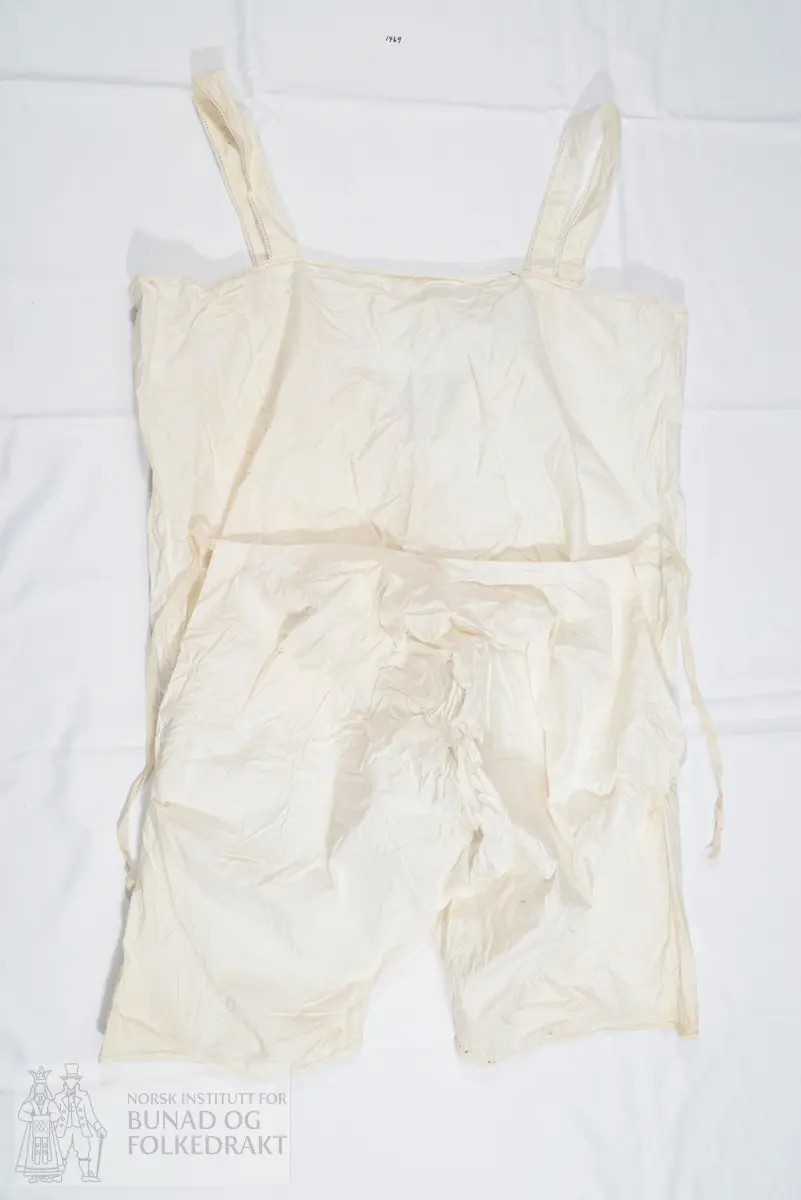 Chemilette, underbukse med overdel/ linnet, i hvit bomull. Knelang bukse, overdel med seler og åpning midt framme med knepping. Splitt i sidene og horisontal åpning bak. Overdelens ryggstykke stanser ved livlinjen. Lokk bak på buksen, med knytesnor øverst på lokket i hvert hjørne. Knytebånd til å knyte rundt livet. Pen avslutning på benene, med sydd hullfald og leggsøm, samt en smal heklekant helt ytterst.