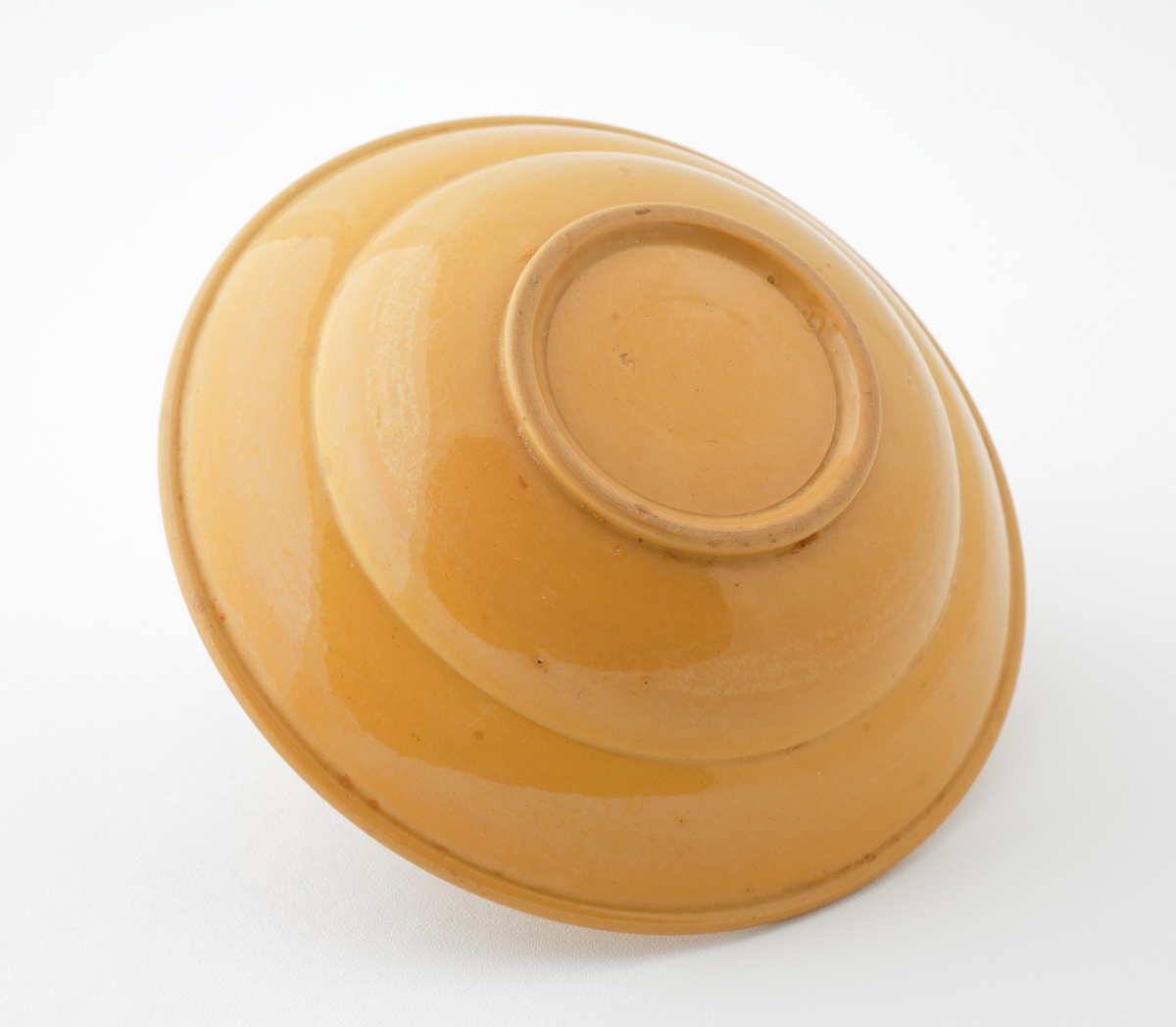 Skål i glaserad keramik.
Rund skål med honungsgul glasyr, försedd med invändig dekorrand (fjäderformationer) i brunt och två gröna ränder i rinningsglasyr. I botten en cirkelrund grön rand.