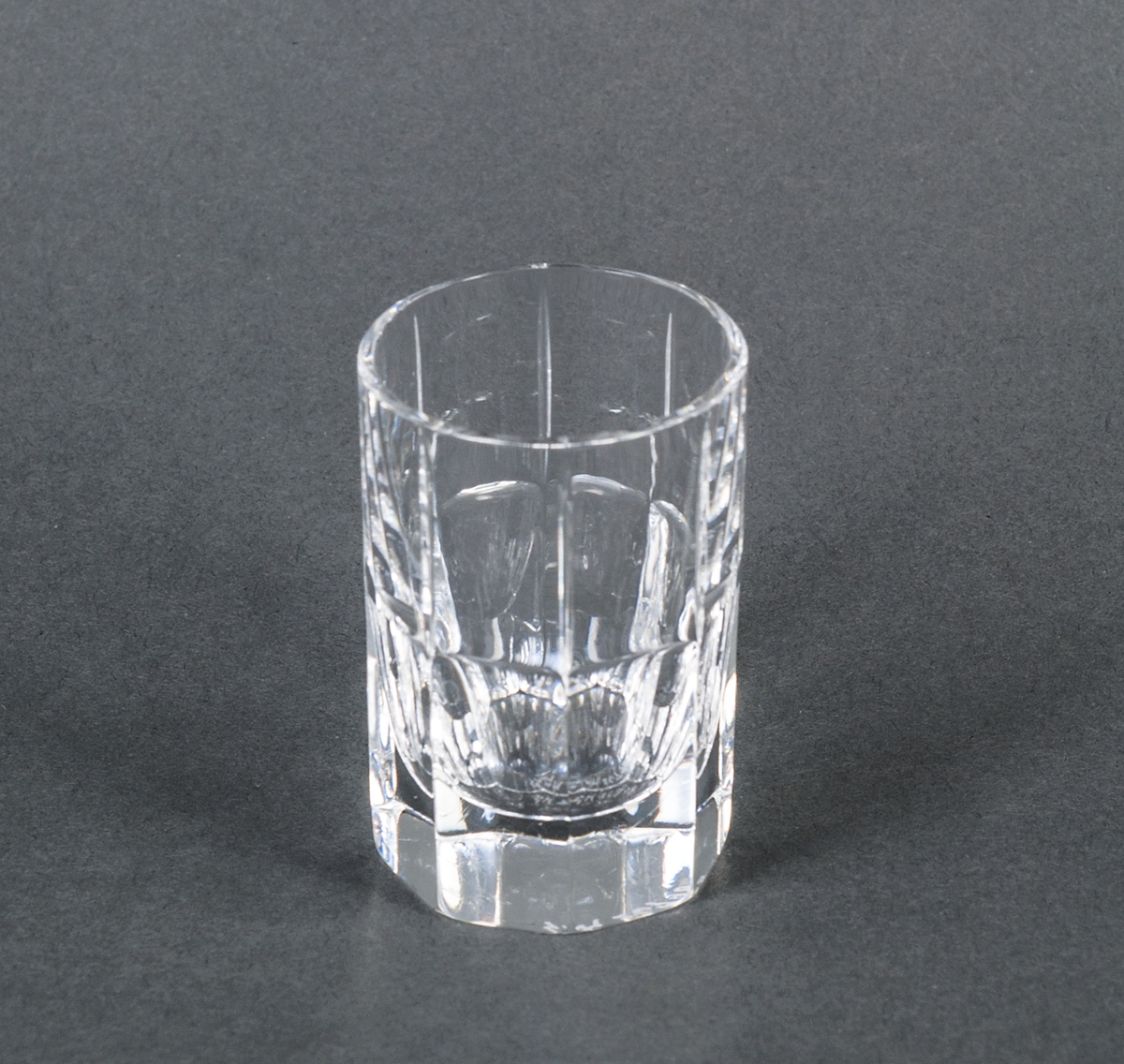 Sake/whiskyglas, ofärgat, prov. Övre halvan cylindrisk med vertikala dekorränder, nedre halvan åttkantig. Design Gunnar Cyrén för Orrefors Glasbruk.