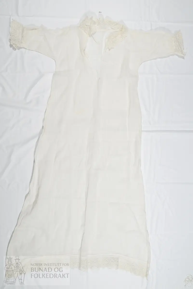 Dåpskjole i tynn hvit bomull, vevd i litt glissen lerretsbinding. Splitt midt bak, lukkes med knytebånd i nakken. Knytebåndet ligger i løpegang i halsen. Løpegang med knytebånd også nederst på ermene. Kragen er laget av kjøpeblonde/maskinprodusert blonde, samme kjøpeblonde er brukt som kanting nederst på skjørtet og nederst på ermene. Blondene er blitt stivet. Ikke tydelig definert liv, sydd med en lang sidesøm. Sidesømmen er sydd i ett med ermesømmen. Maskinsøm med håndsydde detaljer.