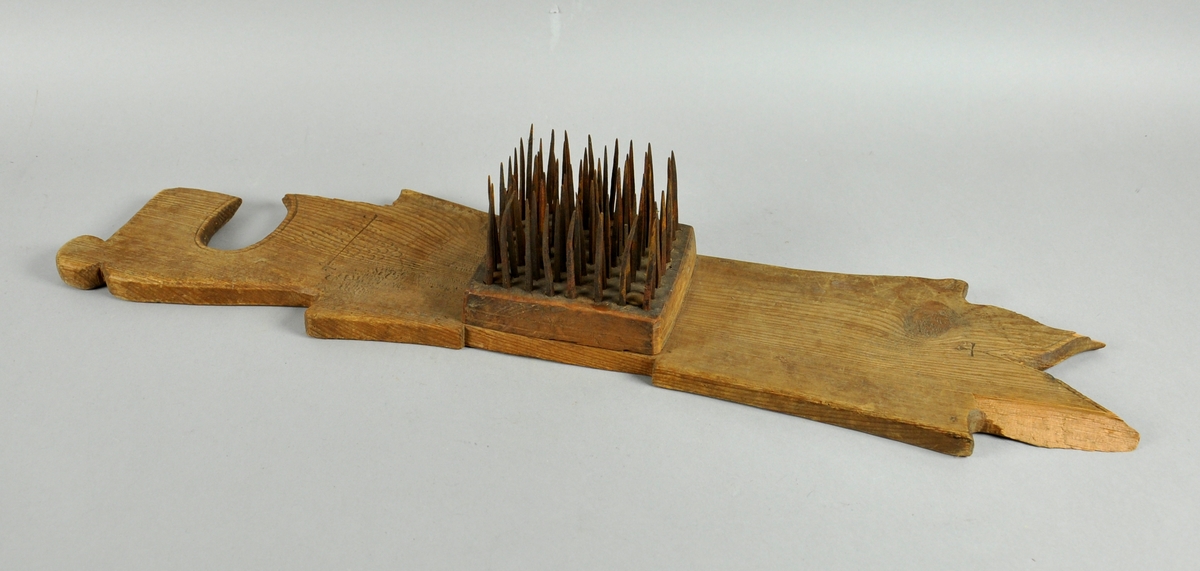 Hampehekle av tre med ovalt hull i den ene enden. Brettet har en trekloss på midten med stålnagler og er skadet i begge ender.