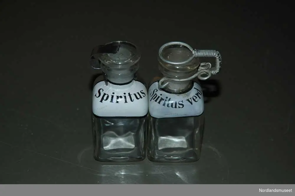 2 gjennomsiktige medisinflasker.
a: Den ene flasken står det "Spiritus" på. Den har en kork på toppen. Den har noe metall greier som holder den på plass.
b: Den andre flasken står det "Spiritus verd." Korken på toppen er feste med en streng rundt.