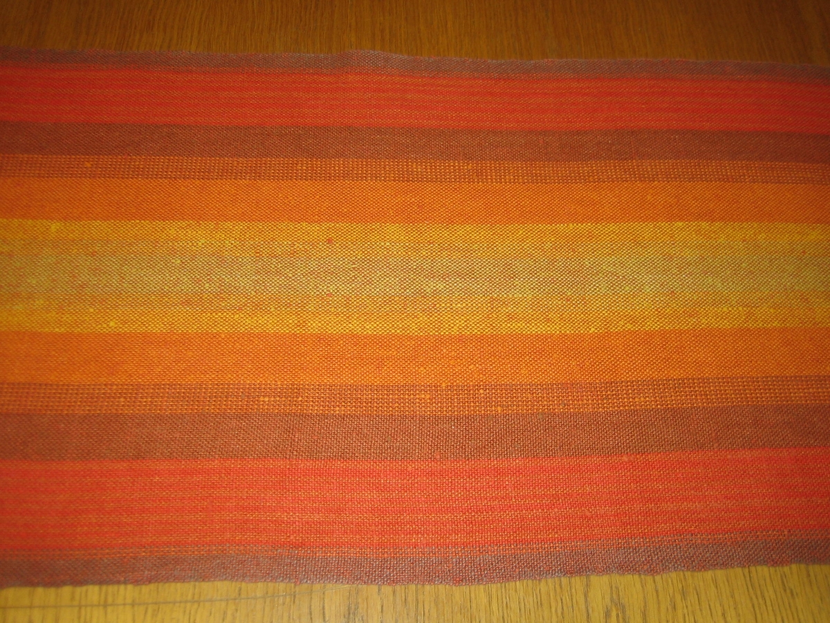 Löpare, vävd i tuskaft för Skaraborgs Hemslöjd i slutet av 1960-talet. Materialet är cottolin i både varp och inslag i flera orange nyanser, brunt och gult.