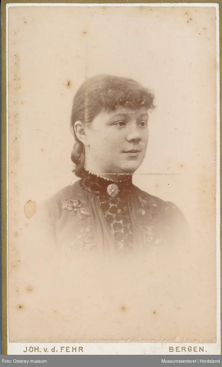 portrettfotografi av ung kvinne med håret samla i nakken og pannelugg. Ho er kledd i kjole/bluse med broderier og mønsta krage og brysstykke, sølje i halsen