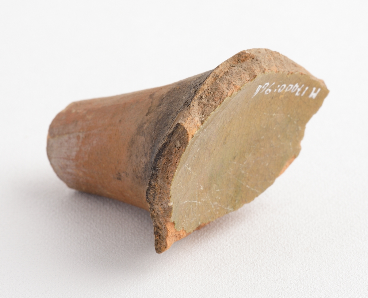 Koniskt ben av yngre rödgods, sannolikt till en trefotsgryta. Benet är avbrutet. Kärlbukens insida har brungul till olivgrön glasyr. Tillverkad i södra Skandinavien.