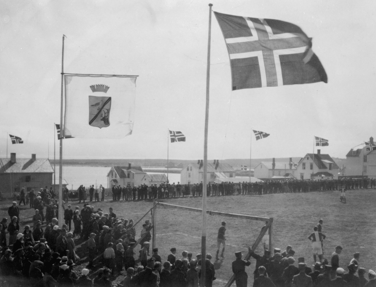 Fotballkamp på idrettsplassen. Flagg og den gamle byvåpen tril Vadsø. En stor tilskuermasse ser på fotballkampen.  Arrangement i forbindelse med 100-års jubileum i 1933.