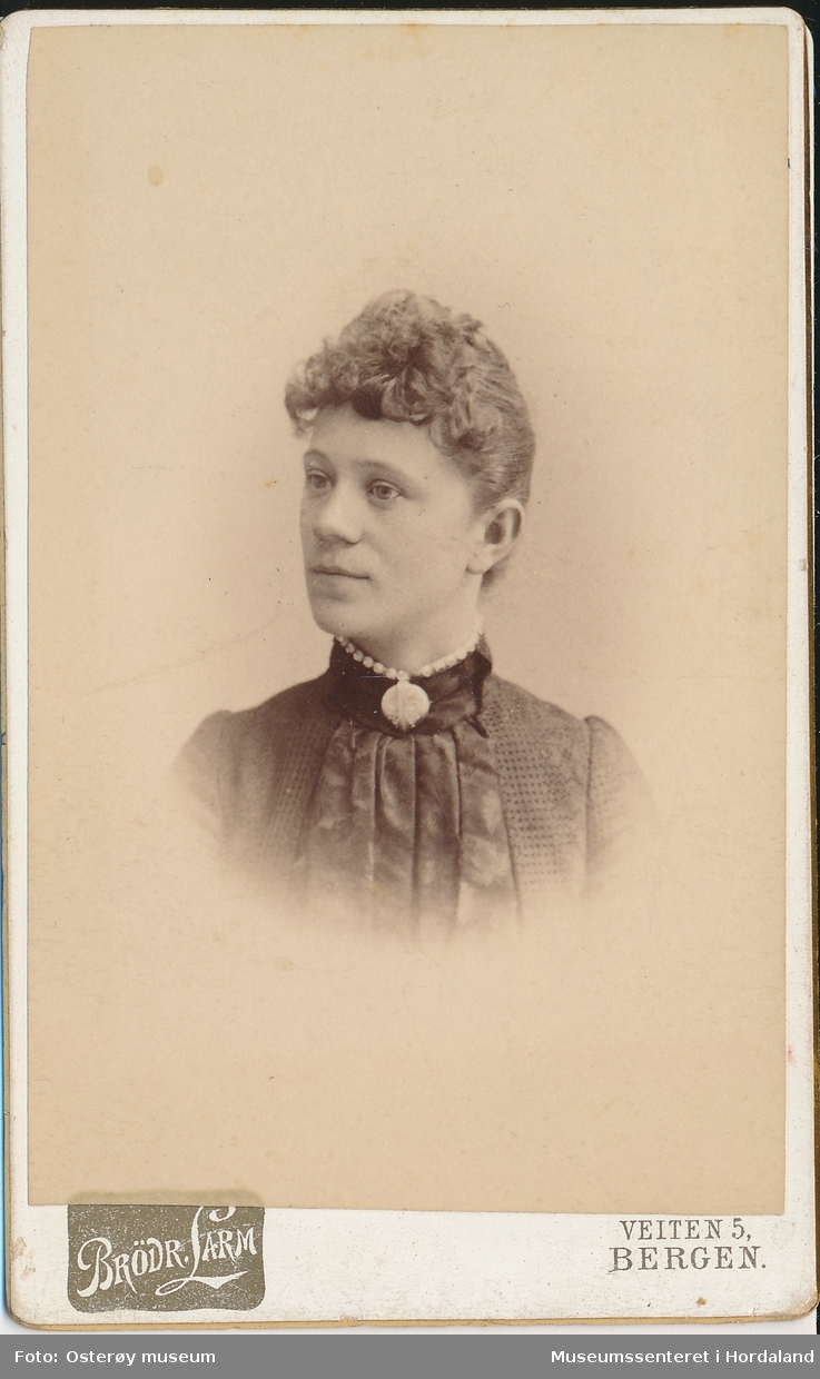 portrettfotografi av kvinne med pannelugg, håret samla i nakken, mørk kjole/bluse med legg og nuperellar og "ponpon" i halsen