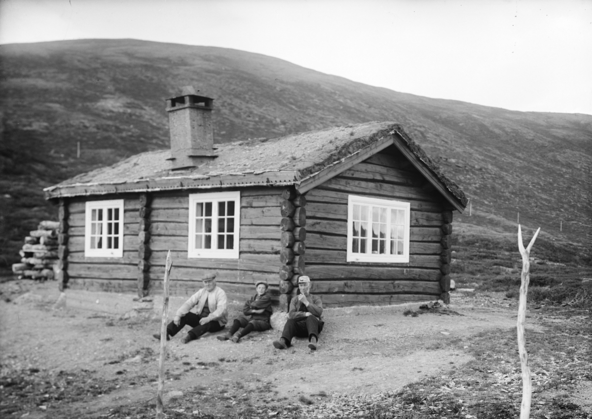 Tømmerhytte på fjellet, to menn og en gutt sitter ved hytteveggen