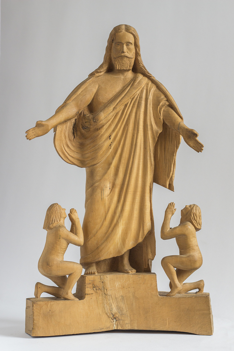 Treskulptur av kristusfigur med to bedende figurer ved føttene.