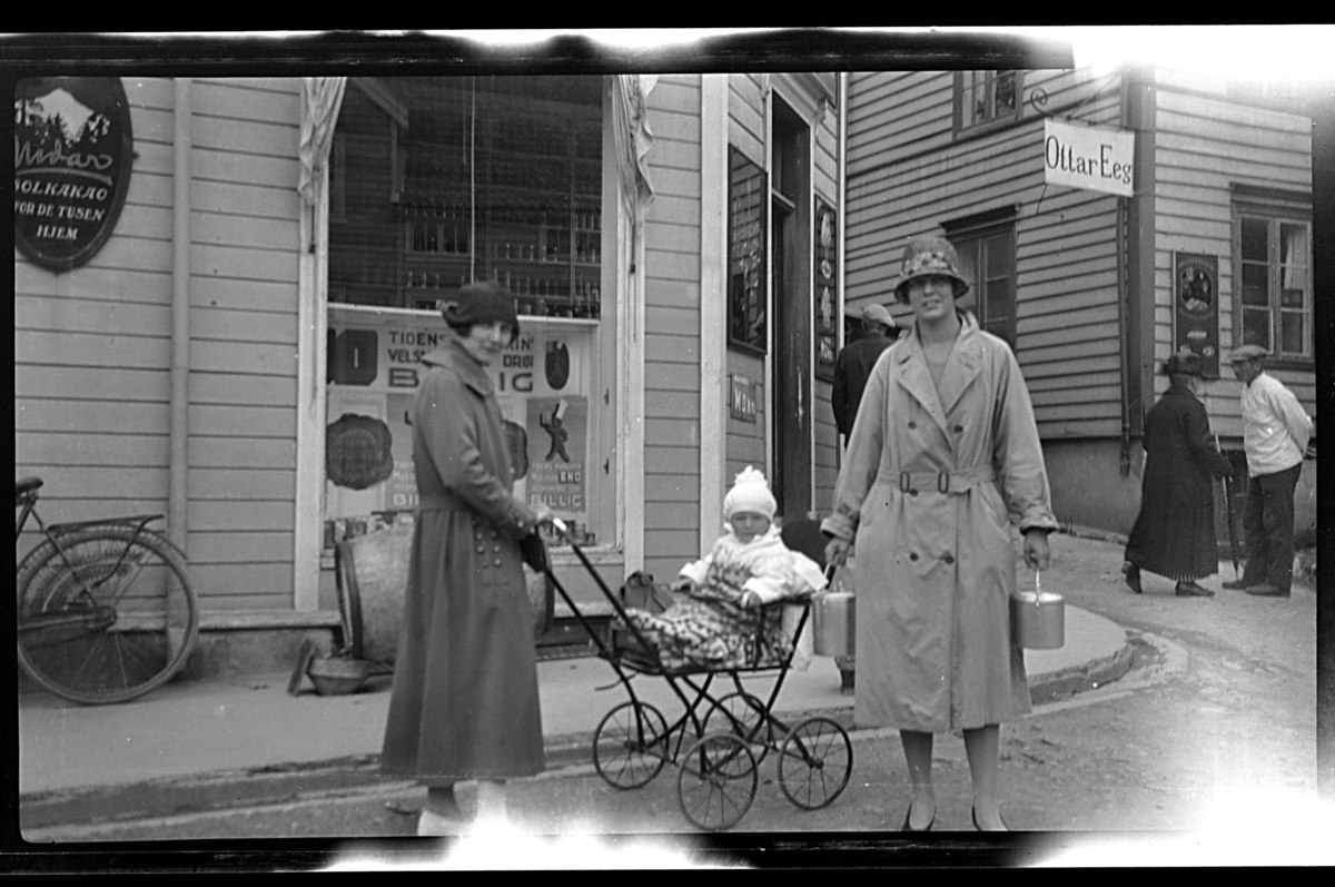 Arne Moestue sitter i en barnevogn mellom sin tante Hilda Sundt og sin mor Lilly Moestue foran en butikk. Fotografert 1926.