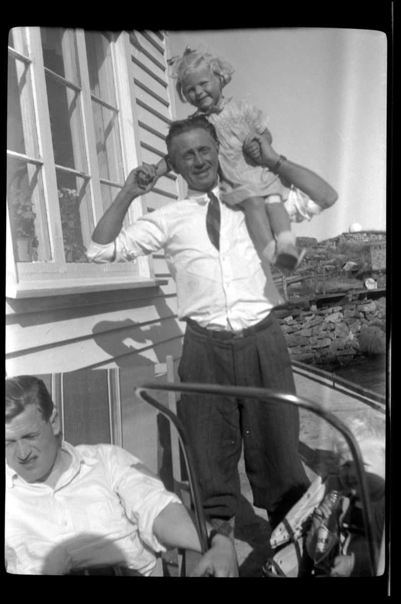 Victor Wilhelmsen bærer en liten jente på skulderen og en ukjent mann sitter i en stol foran Wilhelmsens Sommerpensjonat, Loshavn. Fotografert ca. 1953.