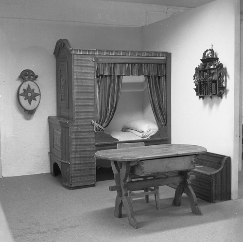 Kämpens i Bollnäs 5 oktober 1982. I ladugården på Kämpens har hembygdsföreningen ett museum där man visar samlingarna. Här ser vi en uppbäddad skåpsäng med omhängen, en vägghylla, samt ett bord och en bänk med förvaringsmöjligheter.
