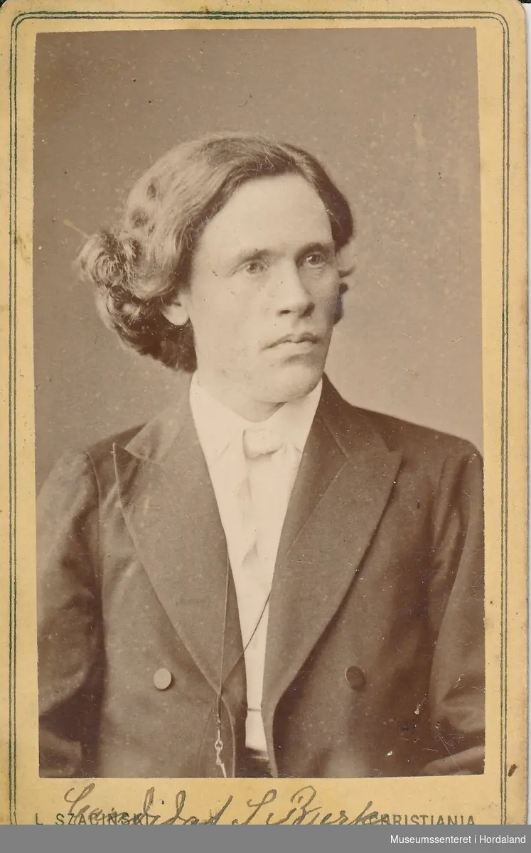 portrettfotografi av mann (brystbilete) med mørk jakke med slag, kvit skjorte og slips med sideksill og halvlangt, bølga hår