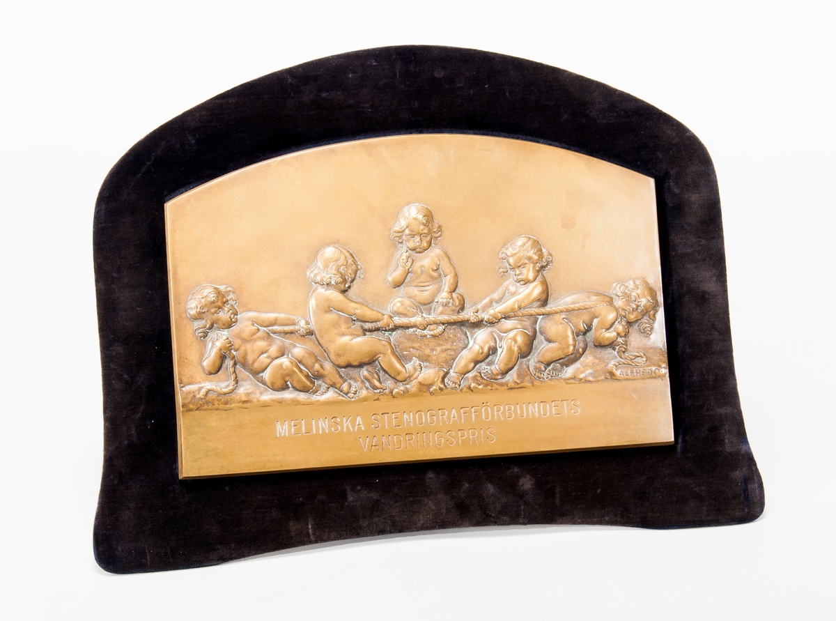 Plakett i brons med motiv av barn som tävlar i dragkamp, med texten "Melinska stenografförbundets vandringspris". Monterad på sammetstavla med stöd för placering på plan yta.