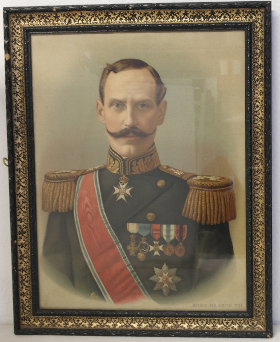 Foto/Trykk av Kong Haakon VII i glass og ramme. Rammen er i svart og forgyllet tre. Den har svarte ornamenter mot en bakgrunn av gull. En lapp fra firmaet som har rammet inn bildet er limt på baksiden av rammen. Oppheng av to små jernringer og et tau.