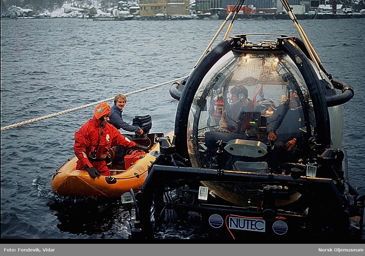 To menn i en liten gummibåt kjører inntil NUTEC's miniubåt Check Mate, som holdes rett over vannoverflaten i Gravdalsbukten. To menn sitter ombord i ubåten. NUI's dykkersenter ligger i bakgrunnen.