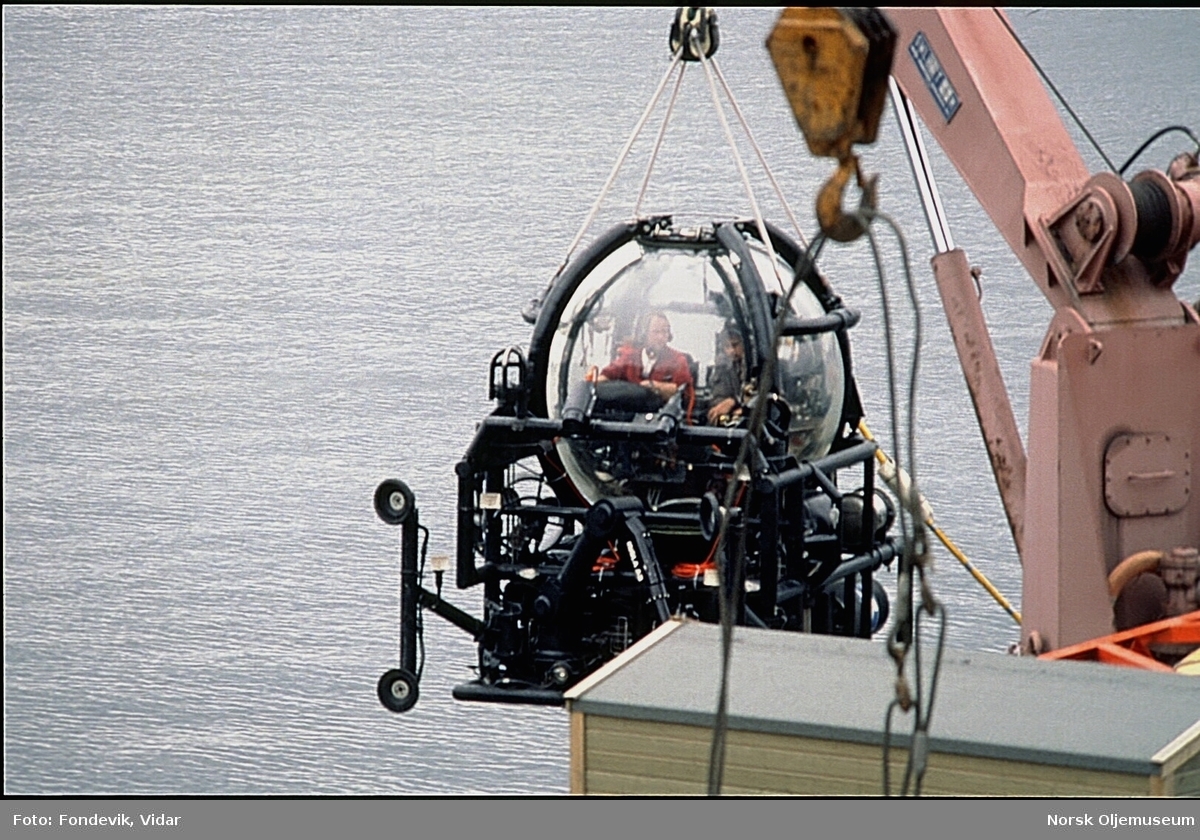 Miniubåten Check Mate til NUTEC holdes over vannoverflaten med en kran på kaien til NUI's dykkesenter i Gravdal. To personer bemanner ubåten.