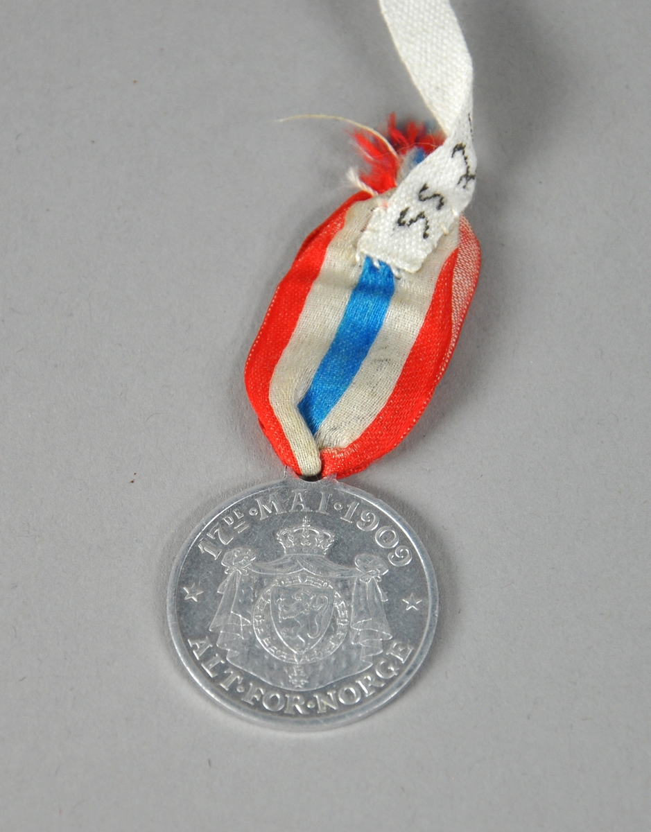 Medalje av aluminium med sløyfe i rødt, hvitt og blått. Medaljen viser portrettene av kong Håkon, dronning Maud og kronprins Olav, med tekst rundt kanten.