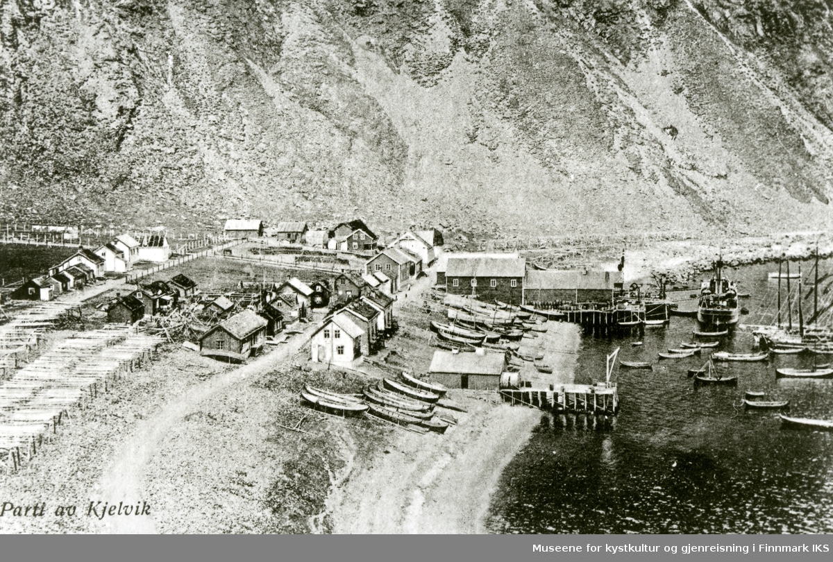 Postkortmotiv. Oversiktsbilde over Kjelvik med bebyggelse, kai, fiskehjeller, båter i havn og Nordlandsbåter dratt opp på stranden. Ca. 1928.