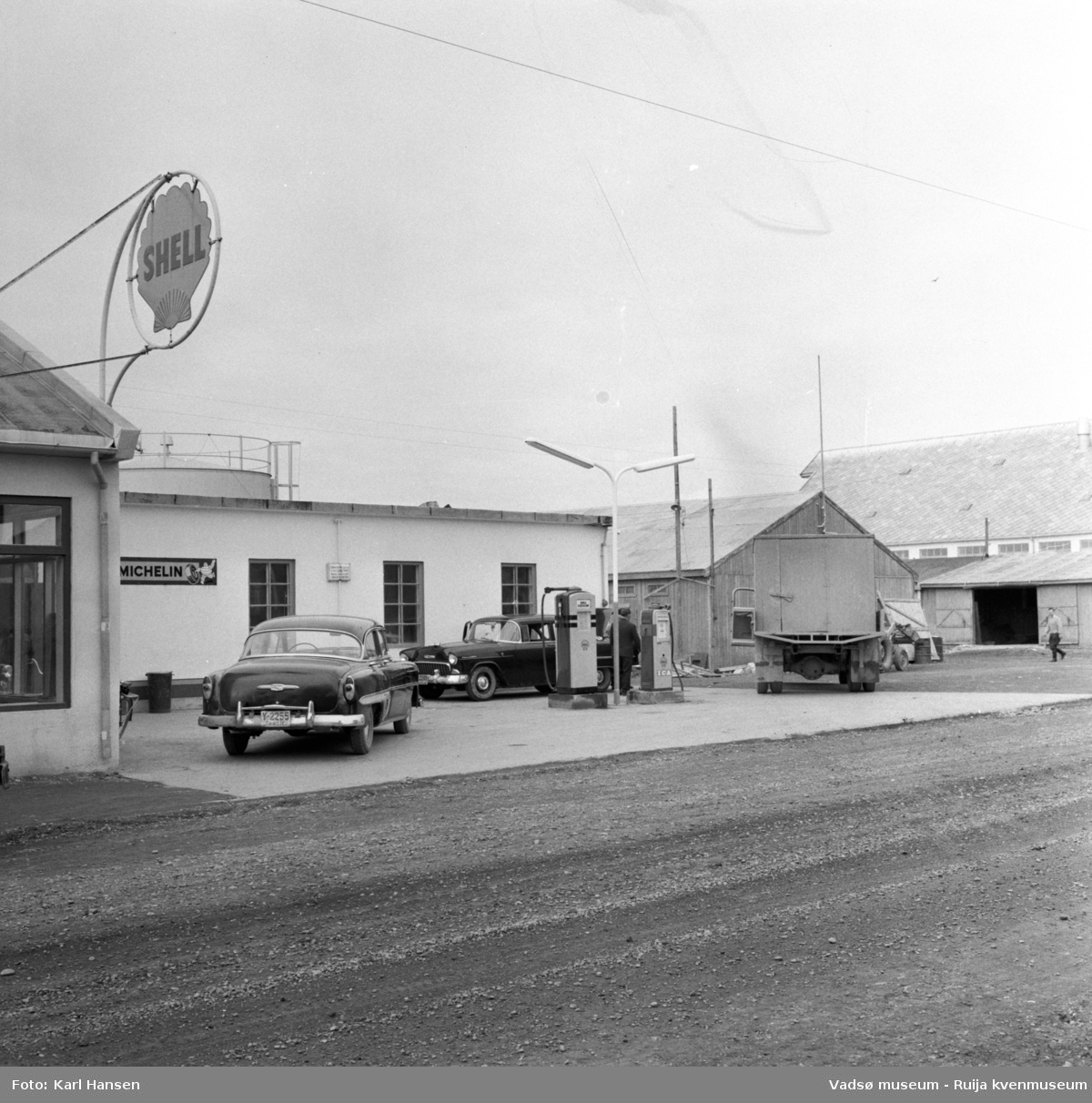 Vadsø sentrum 1959. Shell bensinstasjon og tankanlegg i Tollbugata. Reklameskilt, drivstoffpumper, to drosjer og en lastebil i forkant av bygningen. Til høyre i bildet ser vi skifertaket til Vadsø hermetikkfabrikk.