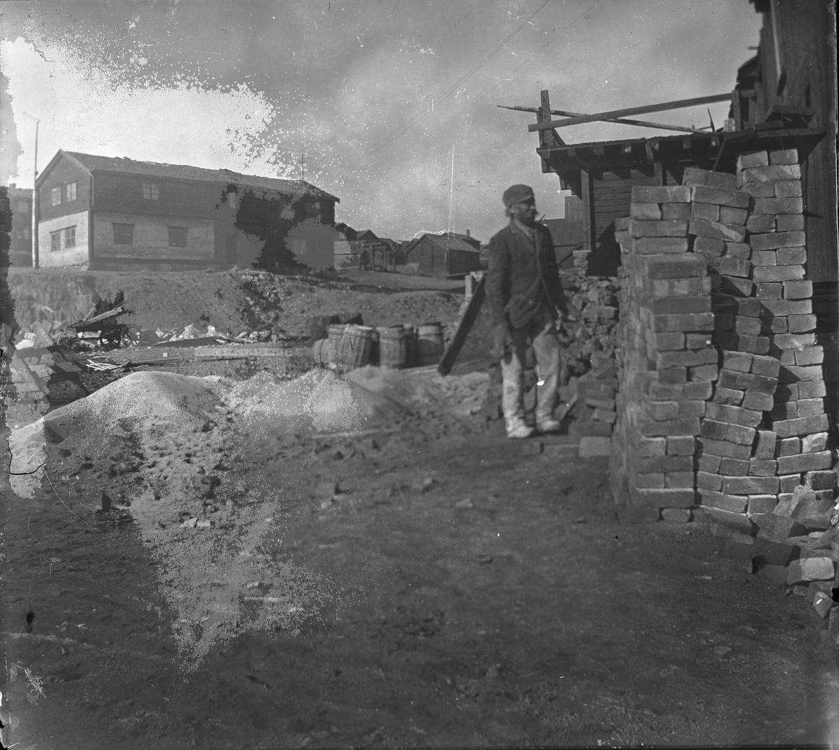 Fra Malmplassen, Røros. En arbeider står ved en stabel med murstein. Hyttstuggu i bakgrunnen