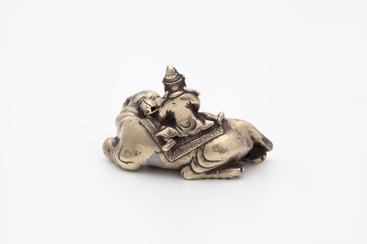 Skulpturen forestiller en mannsfigur som sitters skrevs over ryggen på en liggende elefant. Han er ikledd romslige gevanter og en hatt. Han sitter på et klede med meanderbord og holder i en lotus med begge hendene. Elefanten er dekorert med 6 stjerner.