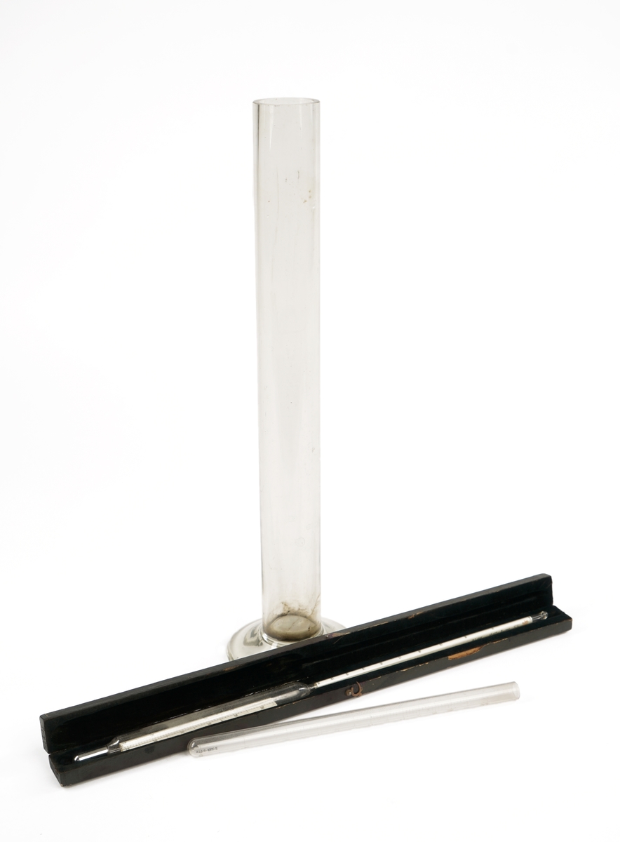 Alkoholmeter/termometer i etui samt glasskolbe og reagensrør. Måleinstrumentet hører ikke til kassen opprinnelig, men fungerer som en erstatning for opprinnelig instrument.