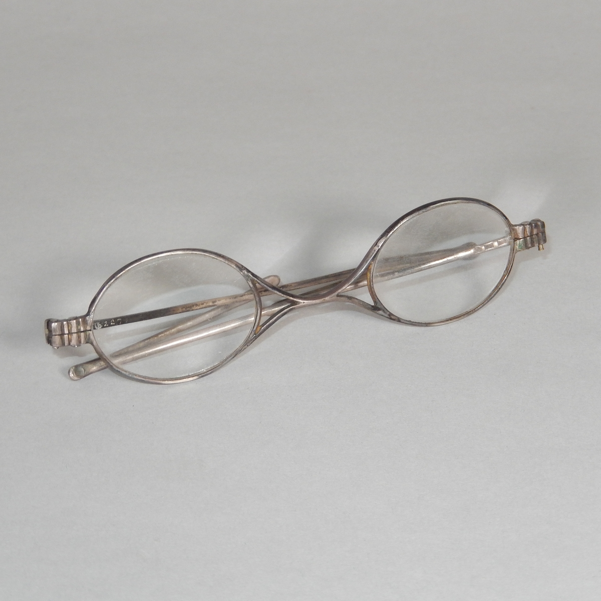 Glasögon med ovala glas och metallbåge. Skalmarna består av två delar som kan fällas ut och böjas. Glasögonfodral, avlångt, av trä med rundade hörn. En del uppfällbar, metallskodd, för att kunna ta ut glasögonen.