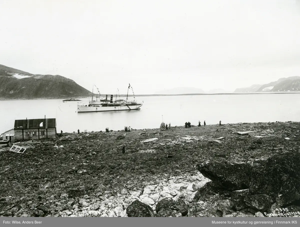 Turister ved Wellmans stasjon i Virgohamna på Svalbard. D/S "Neptun" i bakgrunnen. Til venstre Pikes hus og i midten Andrées støtte. 19.08.1908. (Originalnegativet eies av Norsk Folkemuseum, W 09335)