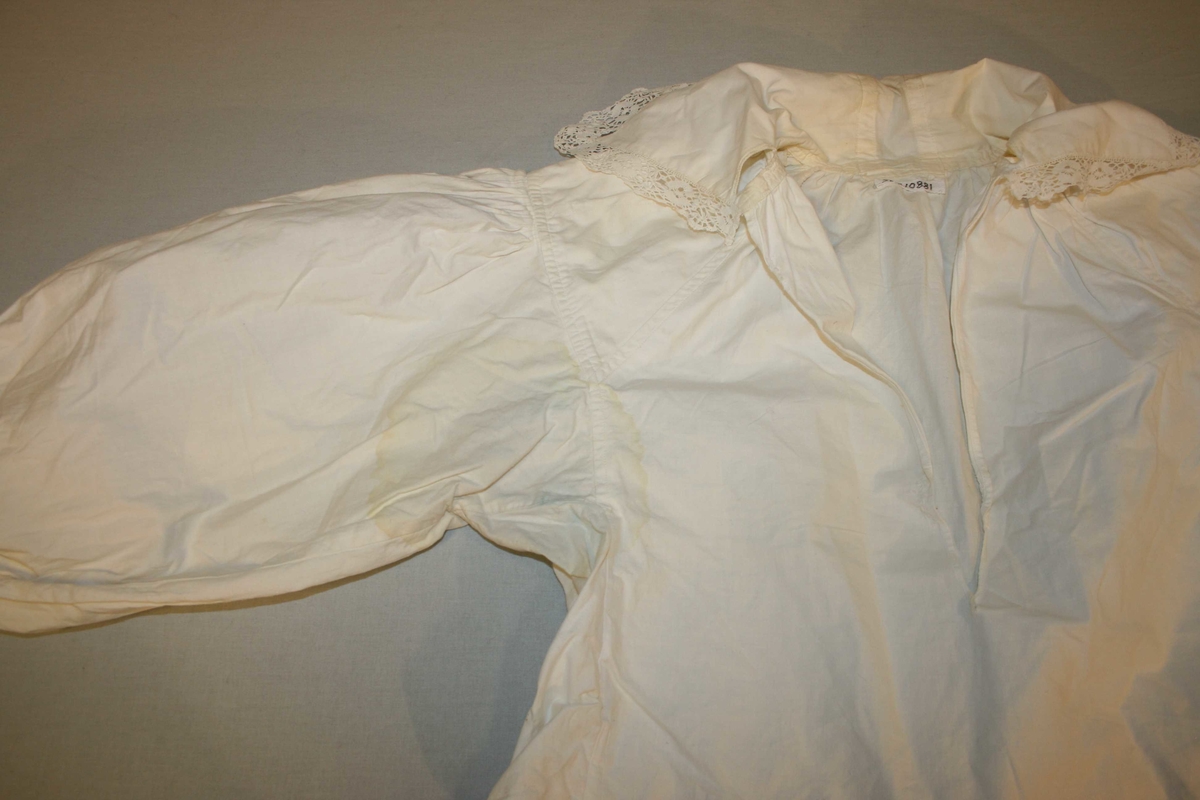 Kvit bomullsskjorte i tradisjonell form. Lange ermar, blonder på krage og mansjettar. Gul under ermane.