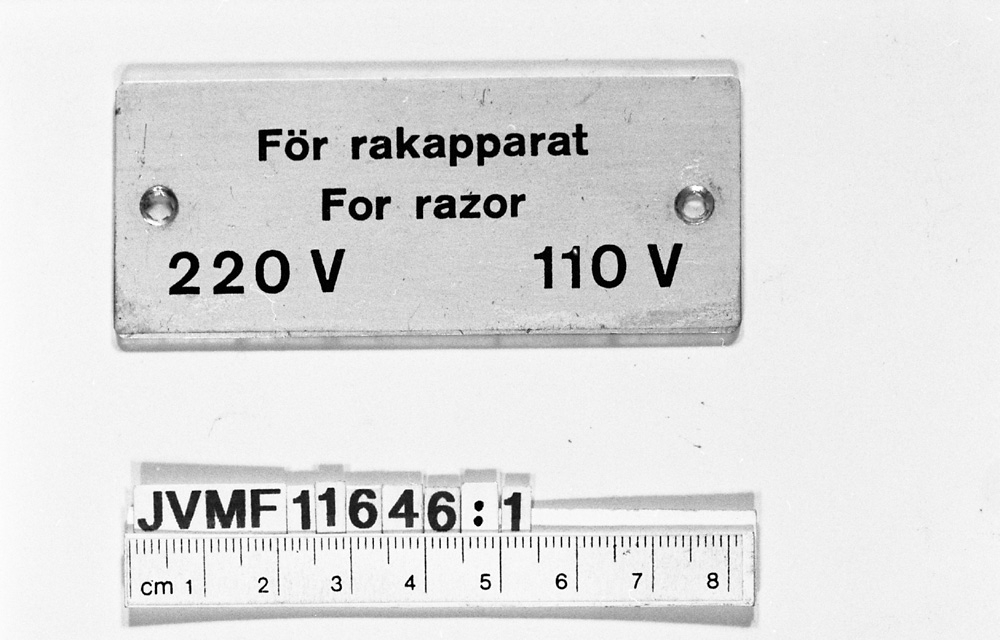 Rektangulär aluminiumsskylt med svart, försänkt text på svenska och tyska:
För rakapparat
For razor
220 V 110 V