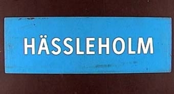 "Rektangulär dubbelsidig plastskylt med vit text på blå botten: 
HÄLSINGBORG C".

På andra sidan:
"HÄSSLEHOLM".