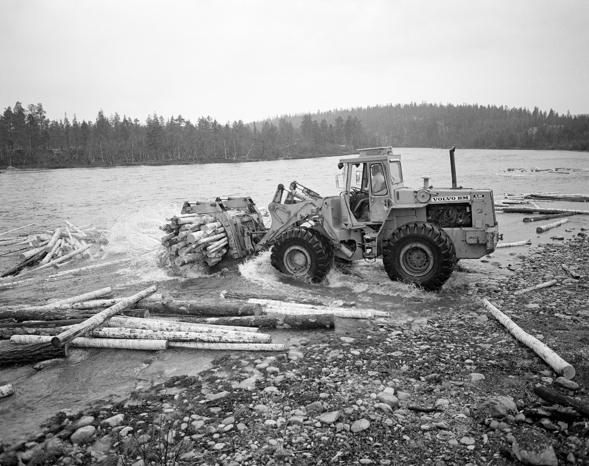 Utislag med hjullaster fra en terminalplass på Røskjota ved Sølenstua i Engerdal i Hedmark. Tømmeret hentes på land, der det var lagt opp ved bredden av Femundselva (Trysilelva).  Med den digre, frontmonterte, hydrauliske kloa som arbeidsredskap hentet maskinføreren tømmerstokkene, som han deretter slapp ut i elva med sikte på at det skulle flyte videre med strømmen. Dette fotografiet ble tatt 22. mai 1986.  Tømmerterminalen på Røskjota ble bygd etter at Klarälvens Flottningsörening i 1953-54 bidro til at det ble bygd bilveger mellom Sølenstua og Galten, og opp til Femundsenden.  Dermed kunne tømmer fra Femund tas opp ved Gløta, og virke fra traktene rundt Isteren kunne hentes ved Isterfossen og kjøres til denne velteplassen, hvor stokkene ble lagt opp for tørking og fløting påfølgende år.  Fra 1965 og framover fikk Klarälvens Flottningsförening opparbeidet flere terminalplasser av den typen vi ser på dette fotografiet i Engerdal og Trysil.  Slike løsninger ble mulige etter at skogsbilvegnettet i de to kommunene var blitt såpass stort at man anså det for mulig å avvikle fløtinga i de minste sidevassdragene. Omlegginga skjedde i forståelse med Trysilvassdragets skogeierforening.  Terminalplassene hadde ikke bare hjullastere, men i mange tilfeller også barkemaskiner, noe som overflødiggjorde det manuelle arbeidet med barkespader, og dermed forenklet driftsprosessene på hogstfeltene vinterstid.
Hjullasteren som ble brukt da dette fotografiet ble tatt var en Volvo BM LM 1240.  Denne modellen ble produsert i perioden 1972-79.  Den veide 12,1 tonn og kunne ta 7-8 tonn i ett grep.

Under utislaget ved Femundselva i 1986 ble det brukt tre hjullastere som alle tilhørte maskinentreprenøren og rallyføreren Olle Arnesson fra Likenäs i Torsby kommune i Sverige.  Maskinførere var Pinnochio Carlson, Eber Halvardsson og Ove Jonsson.