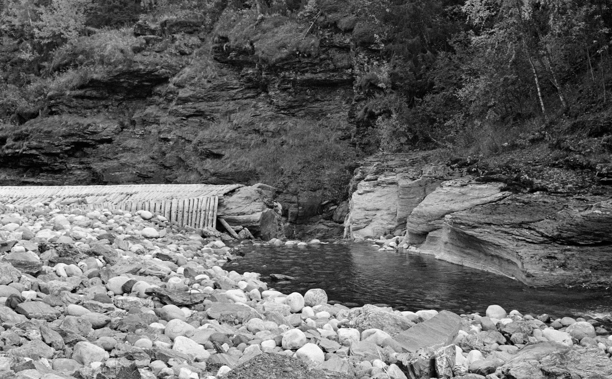 Løftedam ved Loholet i elva Vinstra i grenda Ruste i Nord-Fron, fotografert høsten 1959.  Loholet er en passasje der elva renner i et trangt løp mellom bratte bergvegger.  Her ble det i sin tid bygd en løftedam av tre og steinmasser, som skapte et ni meter høyt fossefall, Vinterbrufossen.  Denne dammen fungerte som vannreservoar for et mølle- og sagbruk like nedenfor, jfr. SJF.1994-02085.  Dette fotografiet er tatt fra dammen, på et tidspunkt da det var sjeldent liten vannføring i elva.  Bildet viser hvorden det vannet som fantes på dette tidspunktet rant ut i et lite skar ved siden av den egentlige dammen.