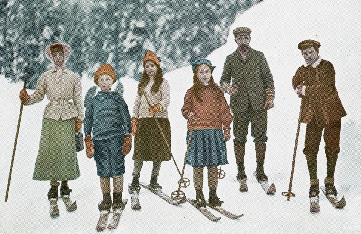 Postkort. Det håndkolorerte fotografiet på kortets fremside viser en mann, en kvinne og fire barn på skitur i vinterlandskap. Skigåing.