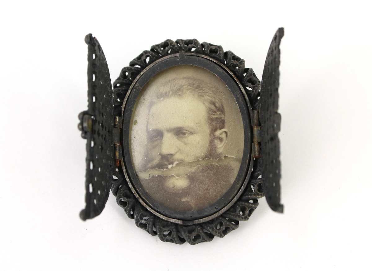 Brosch av metall, oval, framsidan tvådelad som går att öppna och låsa genom en skjutregel. Innehåller ett fotografi av en man.