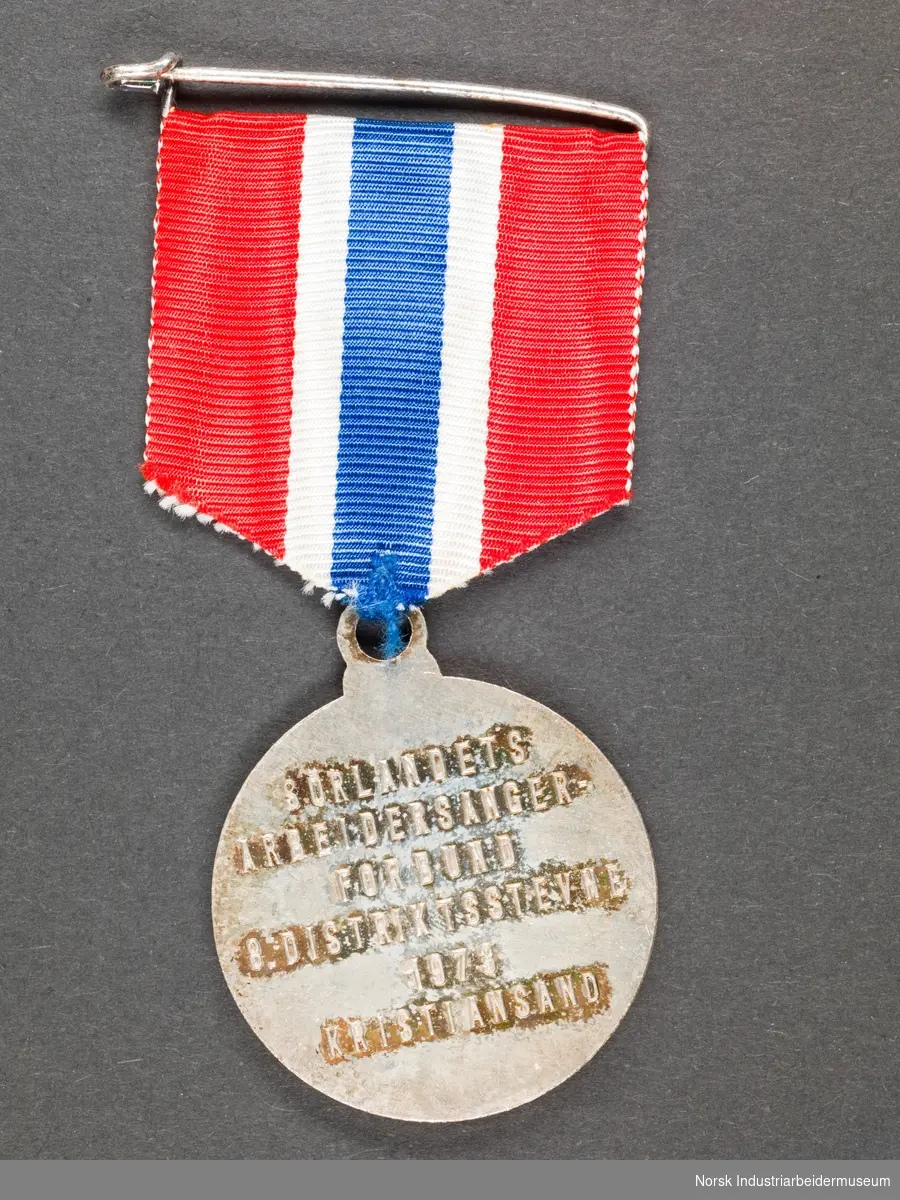 Rund, sølvfarget medalje festet i tekstilbånd i Norges nasjonalfarger. Nål festet til tekstilbånd. Medaljens motiv er på den ene siden en harpe omgitt av en bladkrans og på den andre siden tekst.