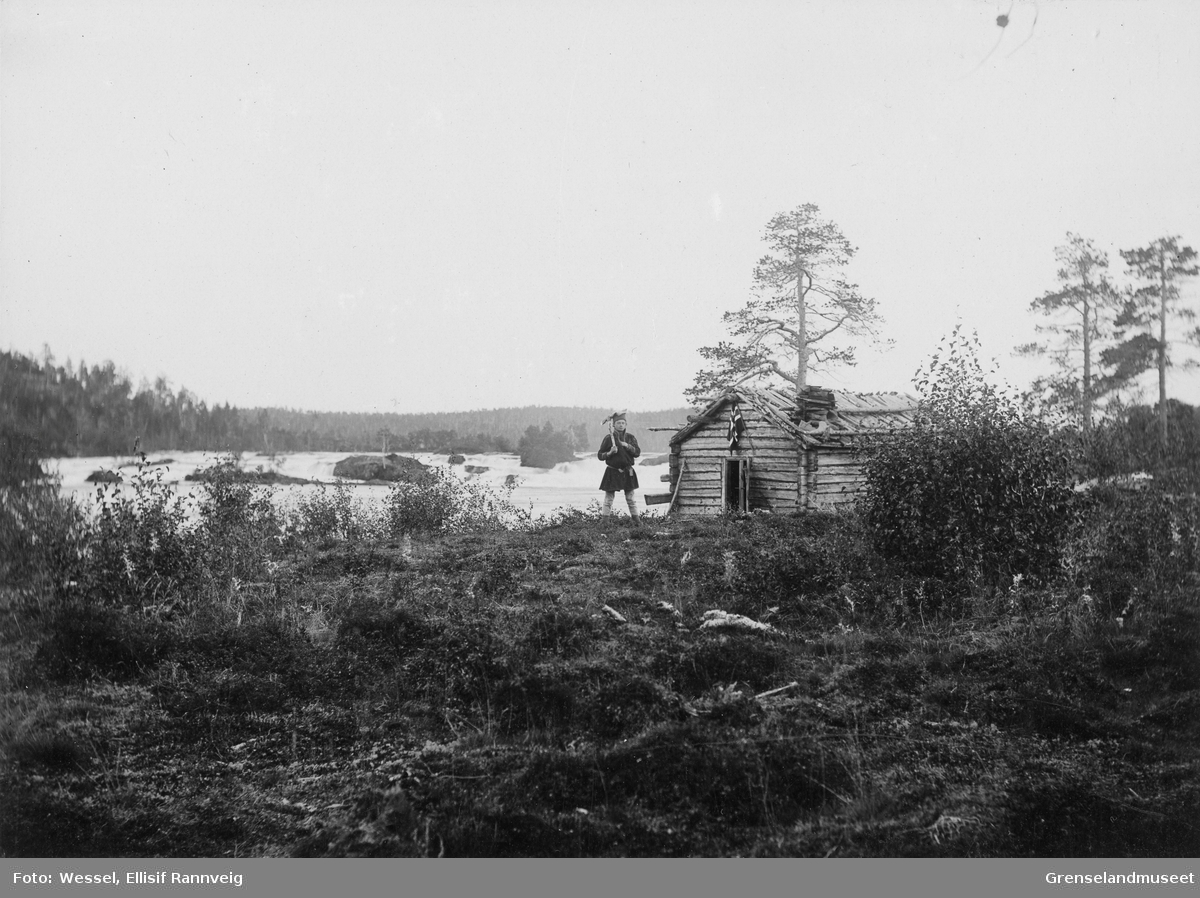 Fra Skogfoss "Männikafoss", september 1896. Ole Johnsen Must står foran koia med en øks over skulderen.
