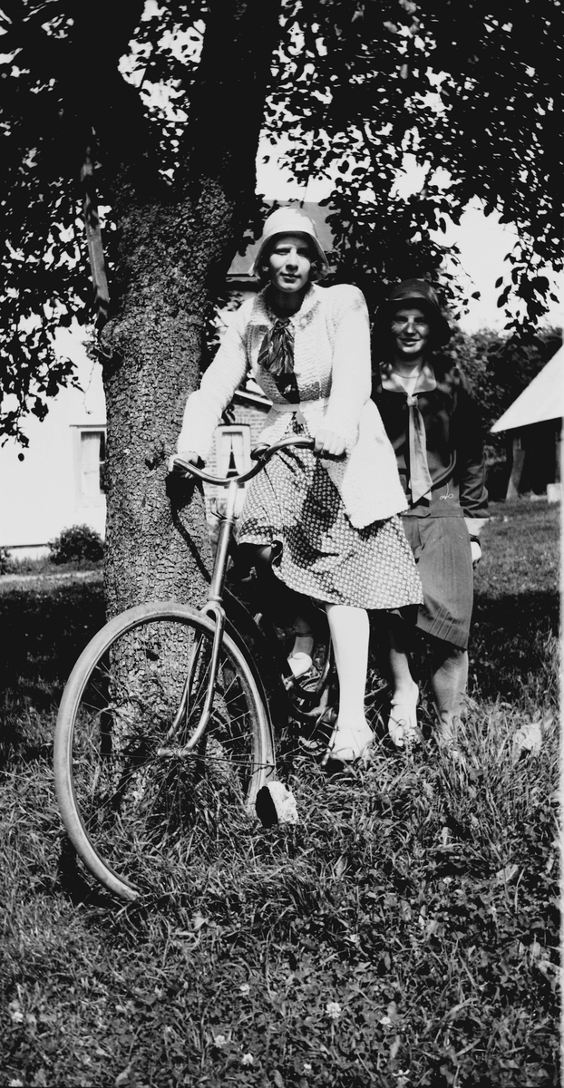 Två yngre kvinnor på en cykel vid ett träd i trädgården. De är uppklädda med klockade hattar, finklänningar och strumpor.  Gårdsbyggnaderna skymtar i bakgrunden.