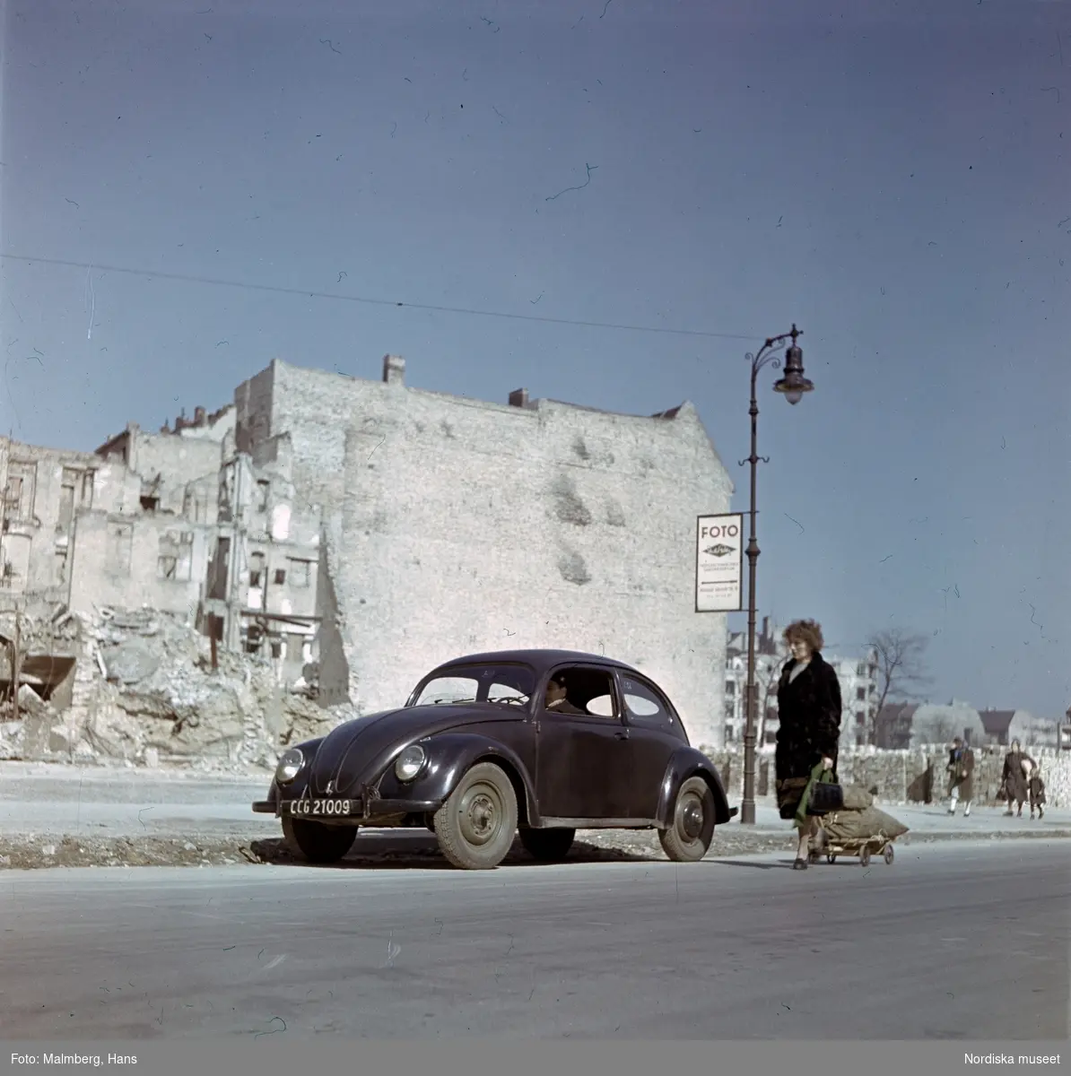 Berlin. På en gata sitter soldat ur de brittiska ockupationstrupperna i en Volkswagen, bredvid drar en kvinna på en kärra.
