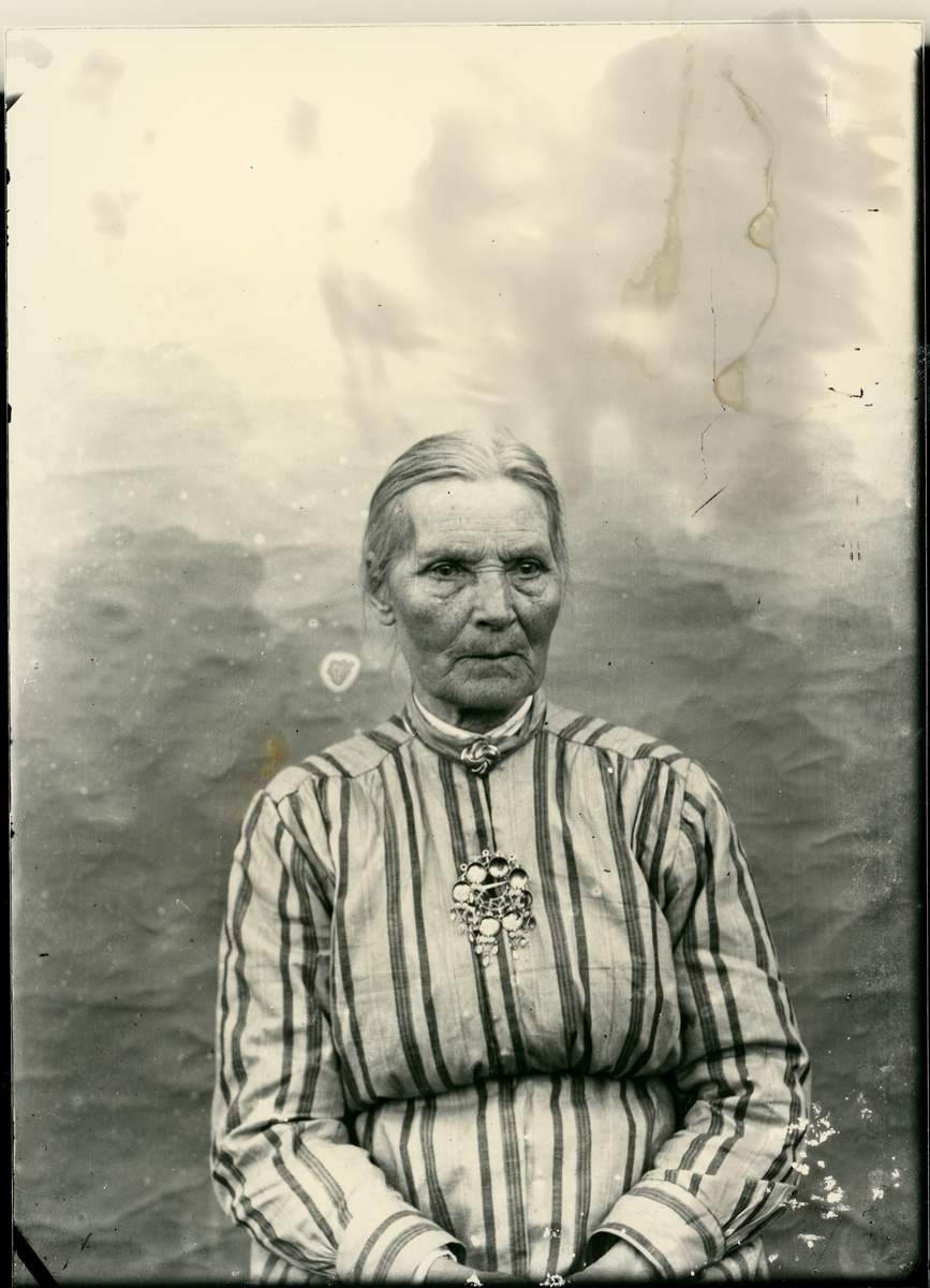Portrett av ei eldre kvinne. Kvinna har oppsatt hår i midtskil og ho har på seg ei stripete kjole med sølje på bringa.