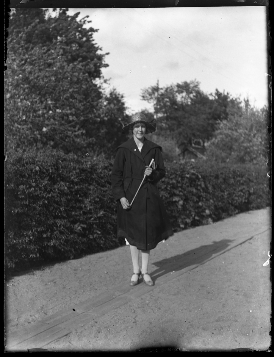 Porträtt av Lydia Landin klädd i svart kappa och svart hatt stående på en grusväg framför buskar. I fotografens katalog såtr "Lydia Landin (Skara)".