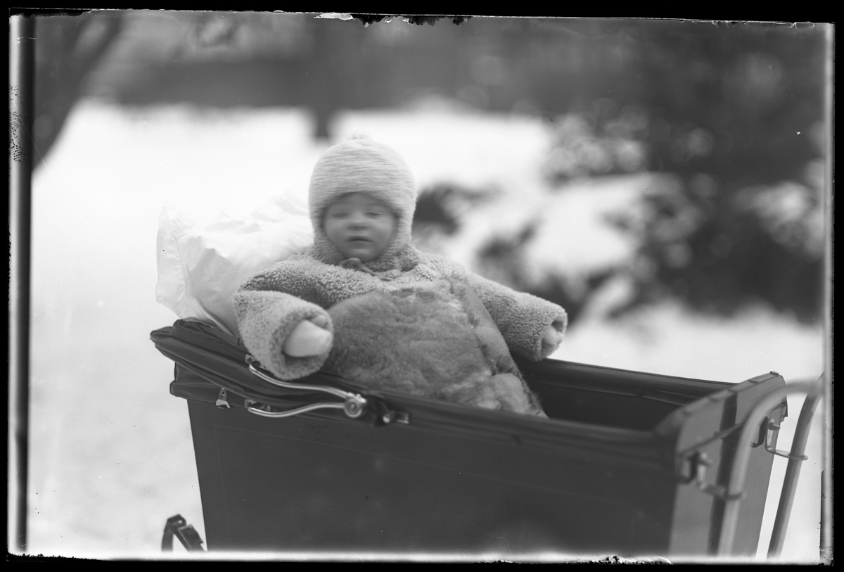 En liten flicka, tolkad som Elsa Margareta Berg, i vinterkläder av päls sitter i en vagn. I fotografens anteckningar står det "Ing[enjör] Bergs flicka i vagnen".