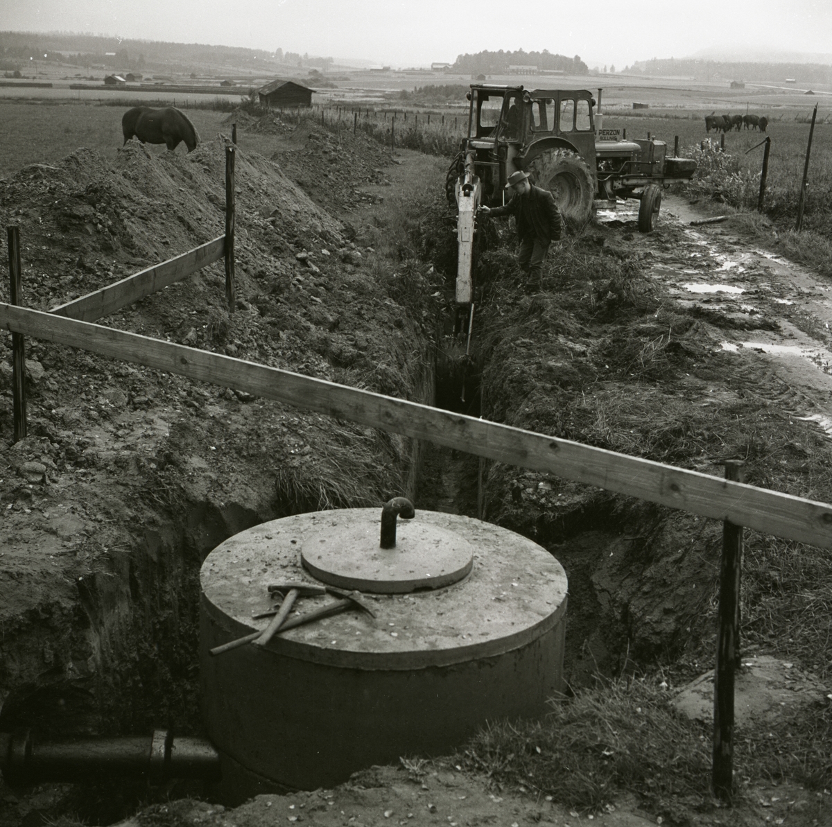 En person gräver med traktor ett dike för nedläggning av rör till avloppsbrunn medan en annan man korrigerar grävarmens riktning, 1967.
