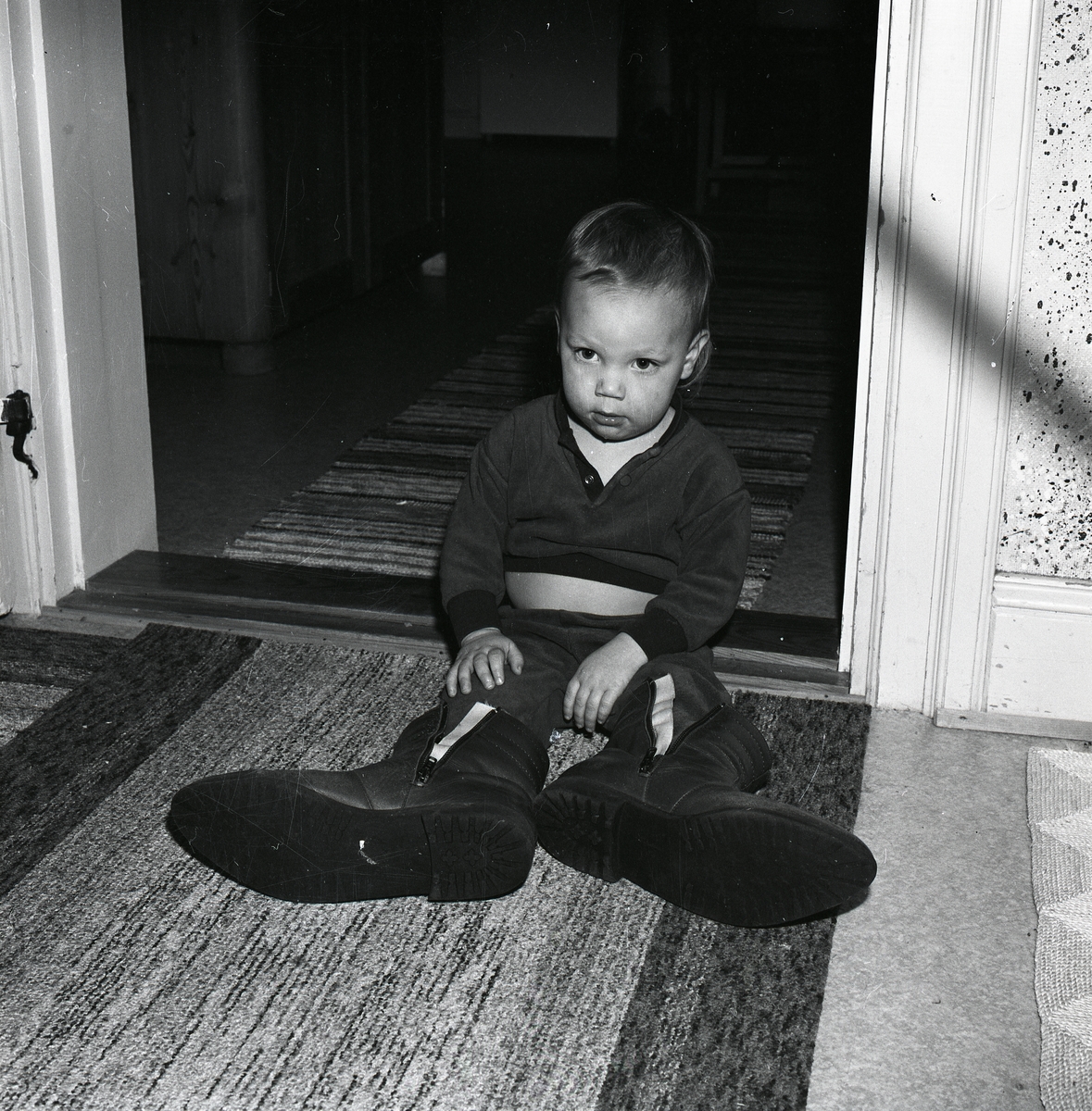 Ett litet barn sitter på en matta på golvet och provar mycket stora skor.