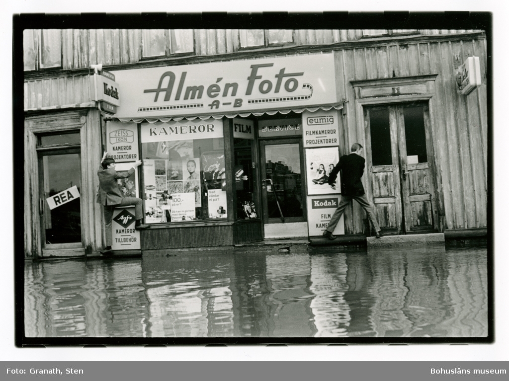 Översvämning utanför butiken "Almén Foto AB". Två unga män i kavaj och kostymbyxor klättrar längs fasaden för att inte gå i vattnet. I och runt skyltfönstret finns reklam för Kodak, Hasselblad, Agfa, Eumig och Zeiss-Ikon.