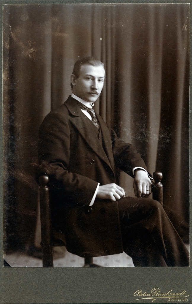 Porträtt av Oskar Träff taget i ateljé. Han sitter i en trebent stol och blickar in i kameran. Han är iklädd tredelad kostym och slips, på vänstra handens ringfinger har han en ring.