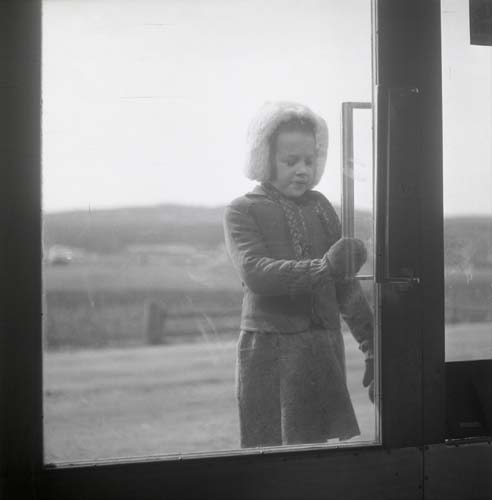 En flicka står utanför en glasdörr med ena handen på handtaget. Hon bär pälsmössa, vantar och tjocktröja, 1949.