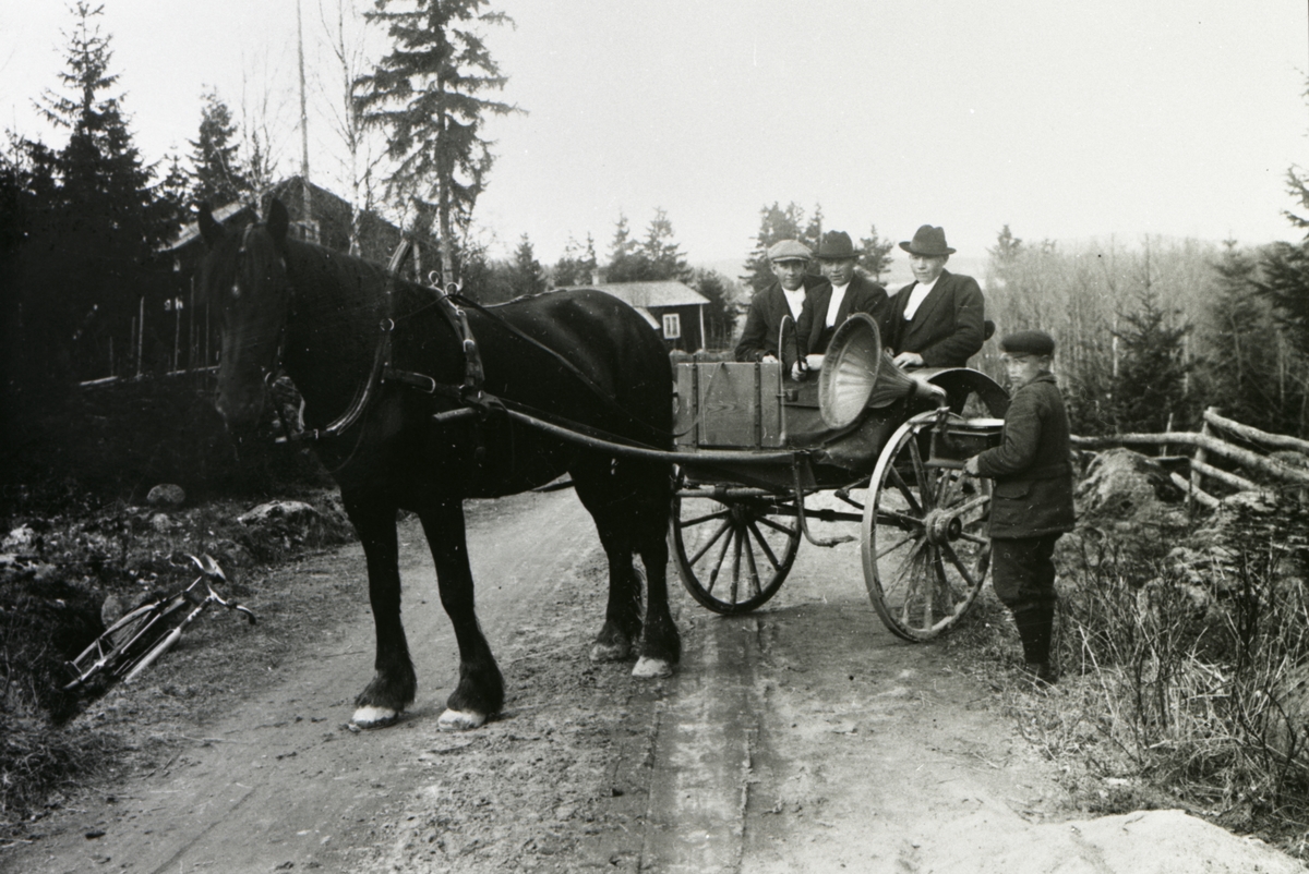Två män och en pojke sitter i en hästdragen vagn medan en pojke står nedanför och håller i en grammofon.