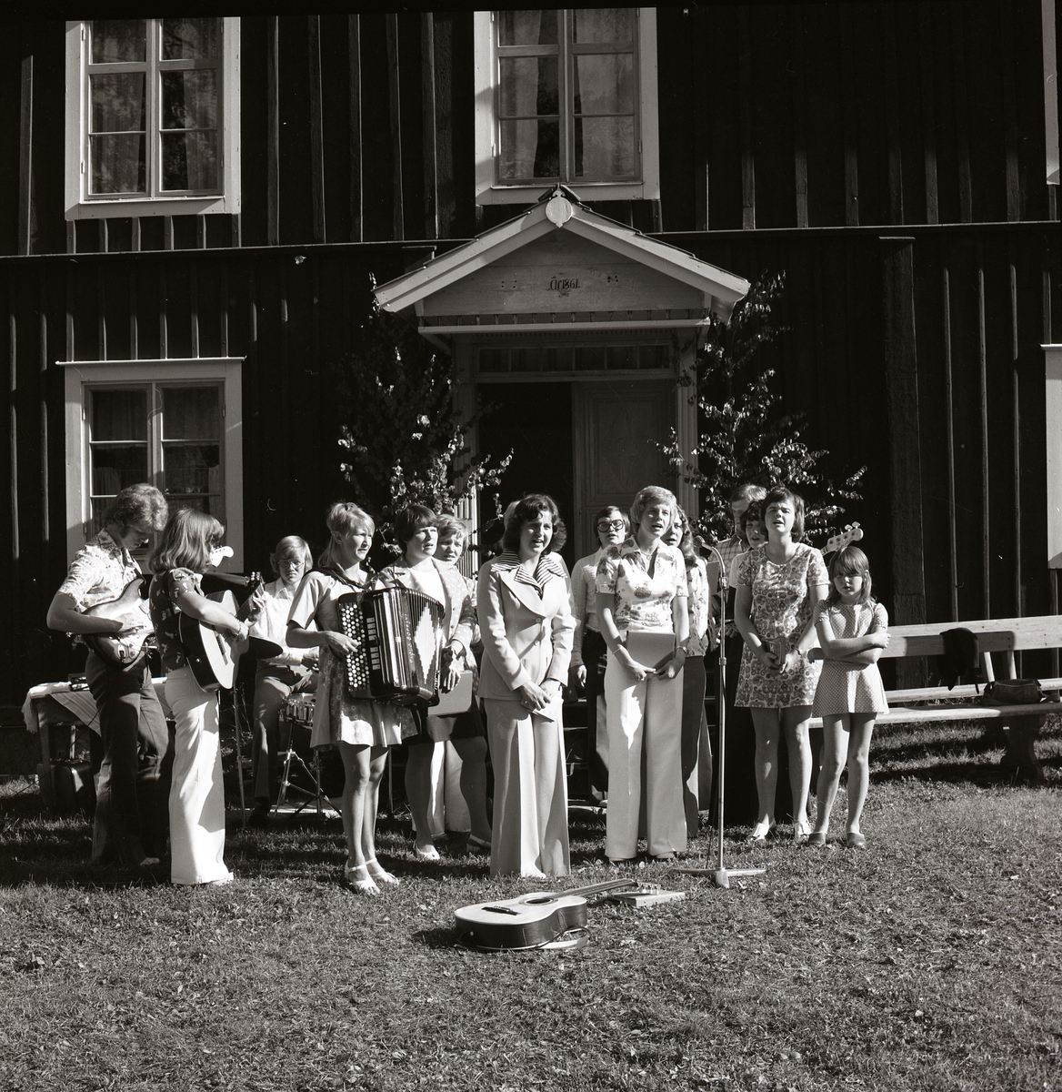 Under en fest vid Rengsjö hembygdsgård sjunger och spelar några kvinnor, män och flickor. Gruppen står framför ett timmerbyggnad och några spelar gitarr och dragspel, 1974.